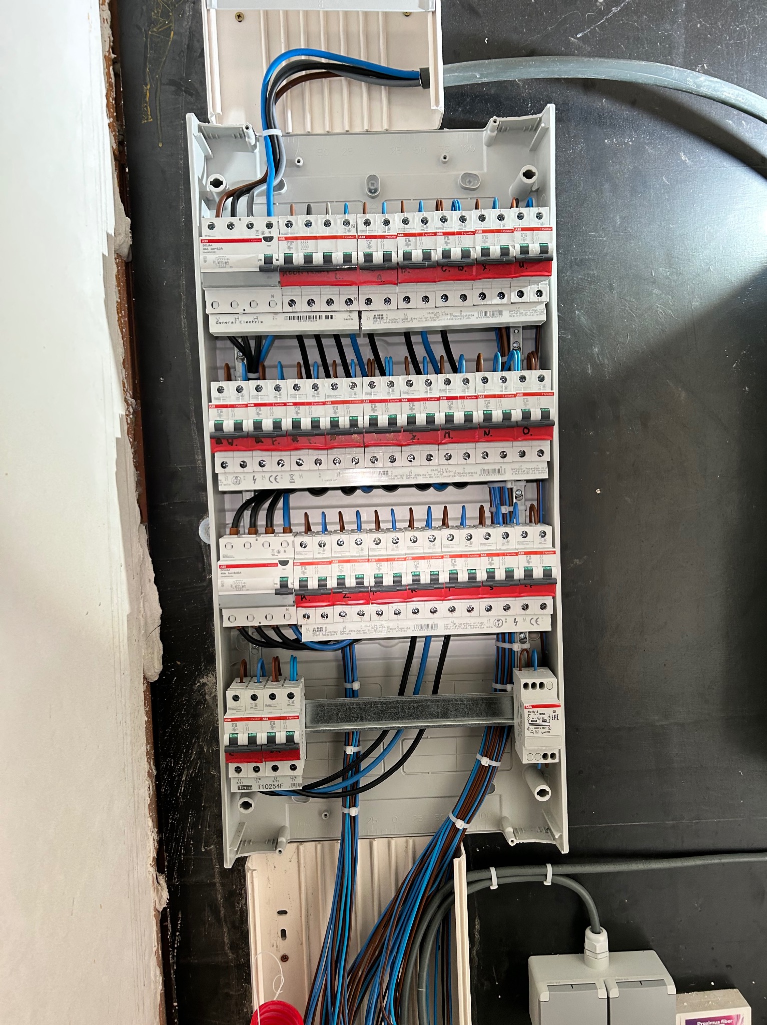 Otwarta skrzynka rozdzielcza z bezpiecznikami, widoczne liczne przewody w kolorach niebieskim, brązowym i czarnym, schludnie ułożone wewnątrz obudowy, zamontowana na ciemnej ścianie obok surowej...