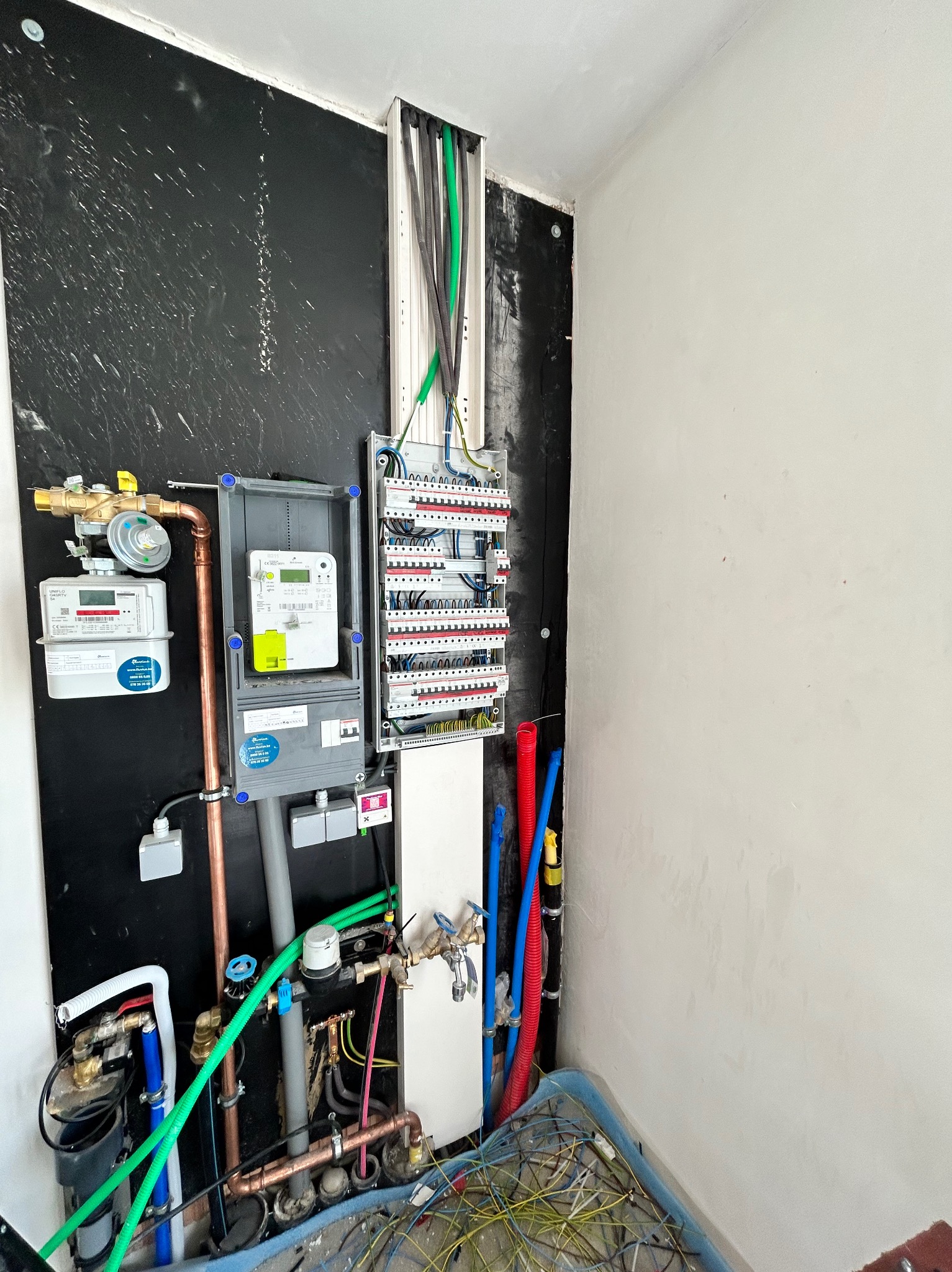 Szara skrzynka z licznikiem elektrycznym obok rozdzielnicy z bezpiecznikami, zamontowane na czarnej ścianie w trakcie instalacji, widoczne przewody i rury instalacyjne w pomieszczeniu w stanie...