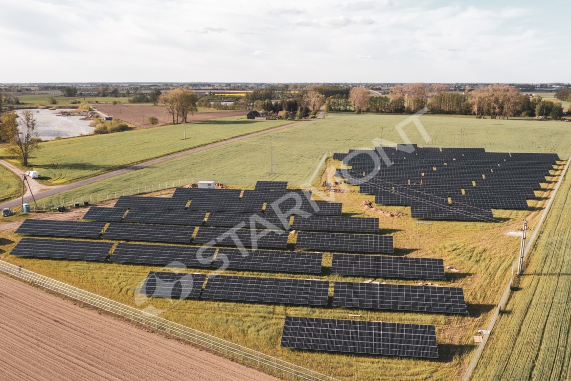 Rozległa farma paneli słonecznych w Leśniewie, widok z lotu ptaka na instalację na polach uprawnych, w tle krajobraz wiejski z jeziorem i zabudowaniami.