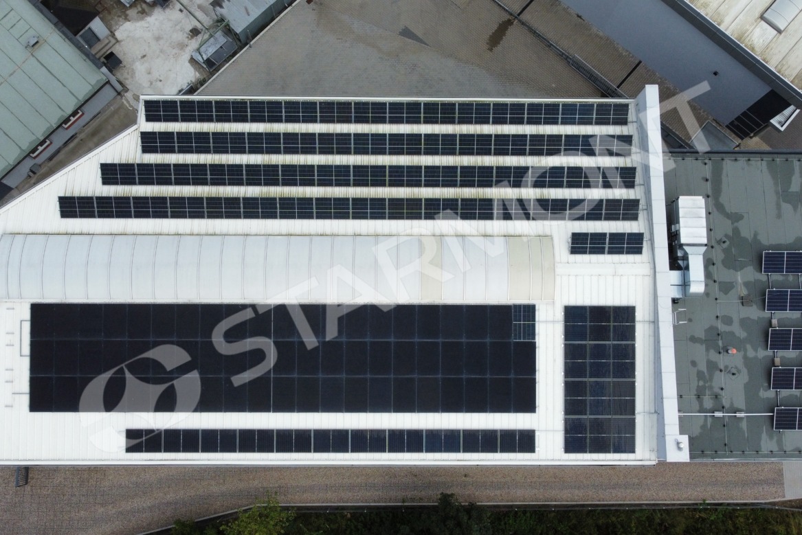 Widok z góry na dużą instalację paneli słonecznych na dachu budynku przemysłowego we Włocławku, z widocznymi różnymi sekcjami paneli ułożonymi w rzędy i otoczeniem innych budynków przemysłowych.