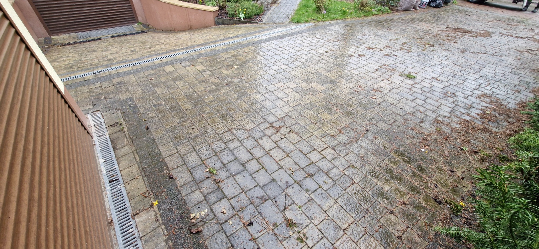 Mokry podjazd z kostki brukowej w odcieniach szarości, z widocznymi liśćmi i igliwiem, przylegający do brązowej ściany budynku z metalowymi żaluzjami i systemem odwadniającym.