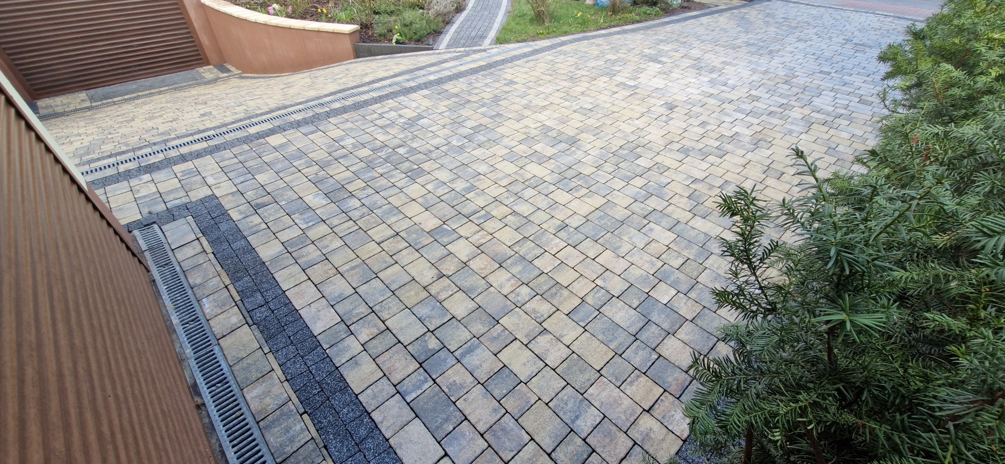 Ułożona kostka brukowa w odcieniach szarości i beżu z liniowymi odpływami wody, widok z góry na posesję z elementami zieleni i fragmentem brązowej elewacji garażu.