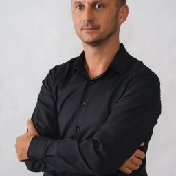 Webiti - Eksperci SEM - Portret mężczyzny w czarnej koszuli z założonymi rękami na jasnoszarym tle.