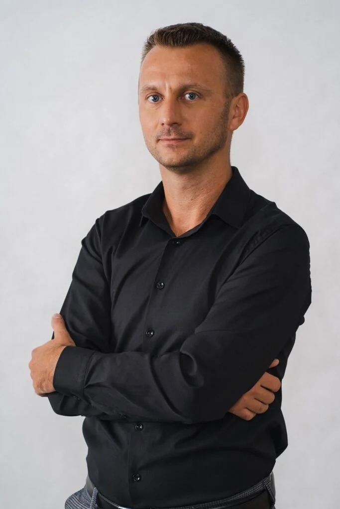 Portret mężczyzny w czarnej koszuli z założonymi rękami na jasnoszarym tle.