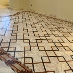 Parquet Artisans - Parkiet z jasnego drewna ułożony we wzór geometryczny z ciemniejszymi akcentami, dekoracyjna listwa przypodłogowa z motywem roślinnym, widoczne listwy maskujące przy ścianie.
