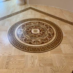 Parquet Artisans - Parkiet z dekoracyjną rozetą i bordiurą w stylu greckim, wykonany w Warszawie. Jasne drewno w kwadratach otacza centralny ornament z ciemniejszych gatunków drewna.