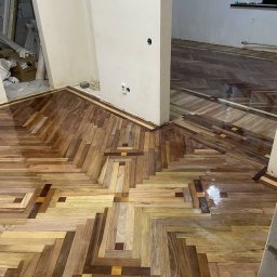 Parquet Artisans - Parkiet ułożony we wzór jodełki z wstawkami z ciemniejszego drewna, częściowo wypolerowany, widoczne gniazdka elektryczne na ścianie.