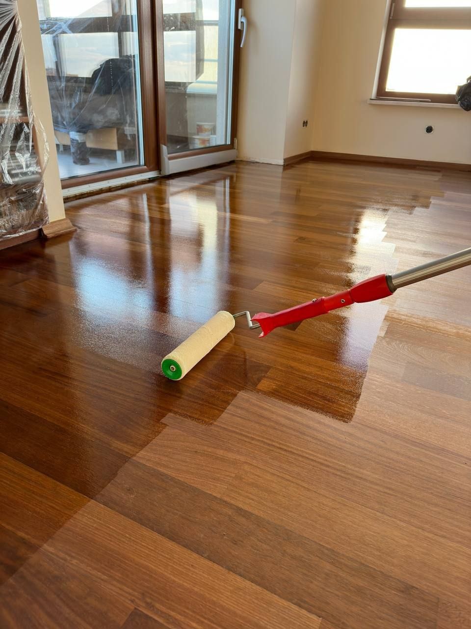 Lakierowanie parkietu wałkiem z czerwoną rączką, widoczny połysk na świeżo polakierowanej podłodze drewnianej w jasnym pomieszczeniu z oknami.