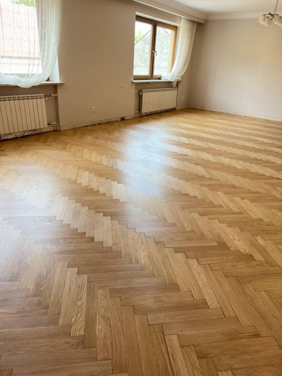 Jasny, odnowiony parkiet ułożony w jodełkę w pustym pokoju z dwoma oknami i grzejnikami.