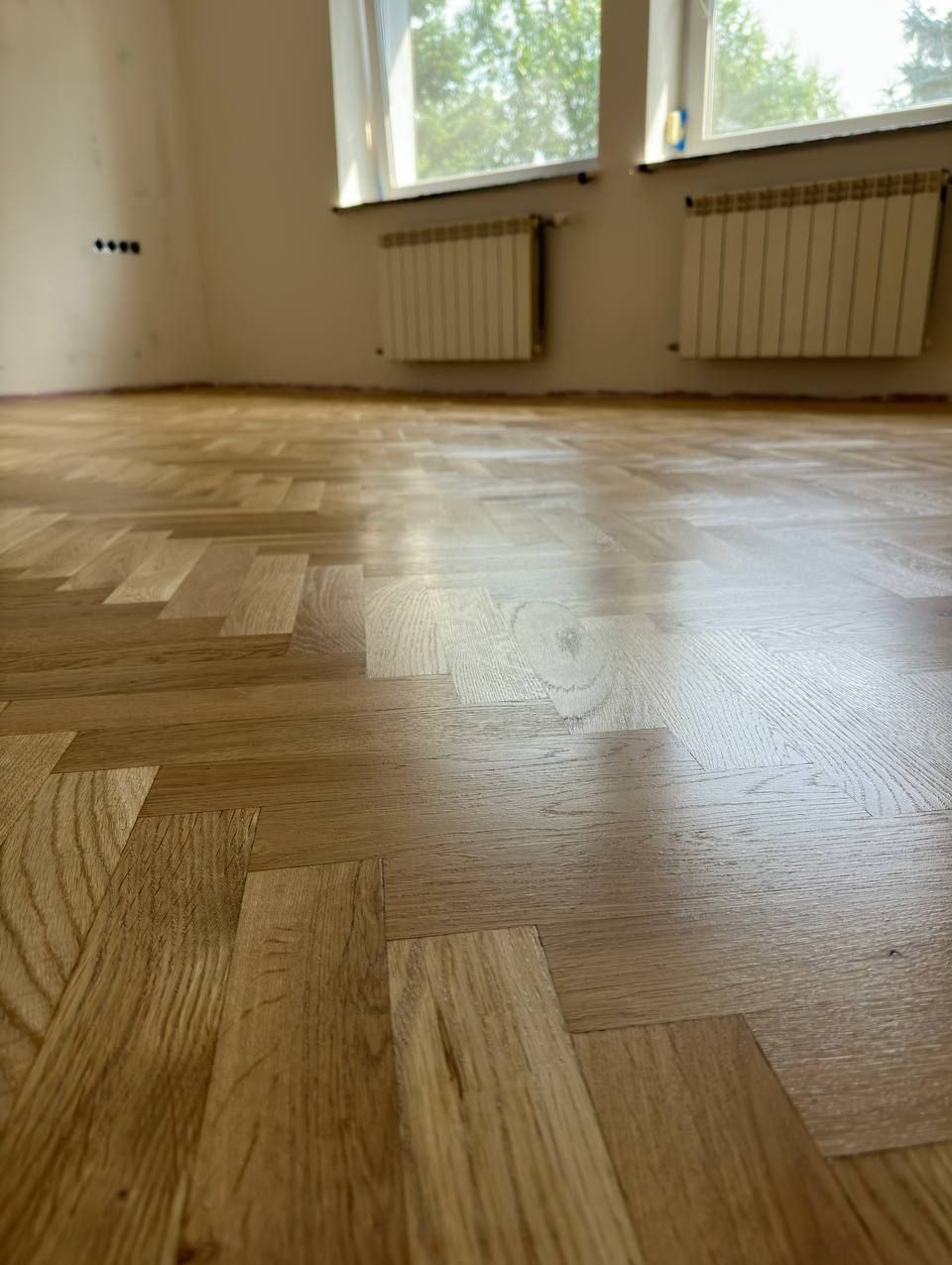 Jasny, lakierowany parkiet ułożony w jodełkę, widok z niskiej perspektywy, ślady po taśmie malarskiej na ścianie, dwa grzejniki pod oknami.