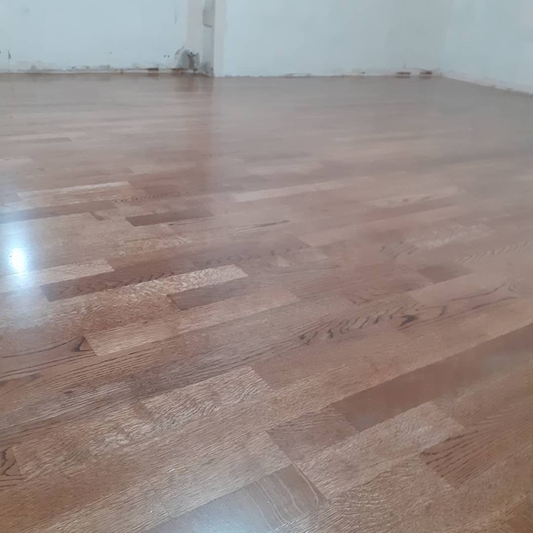 Błyszcząca, wycyklinowana podłoga parkietowa z naturalnego drewna w pustym pomieszczeniu, widoczne drobne niedoskonałości na ścianie w tle.