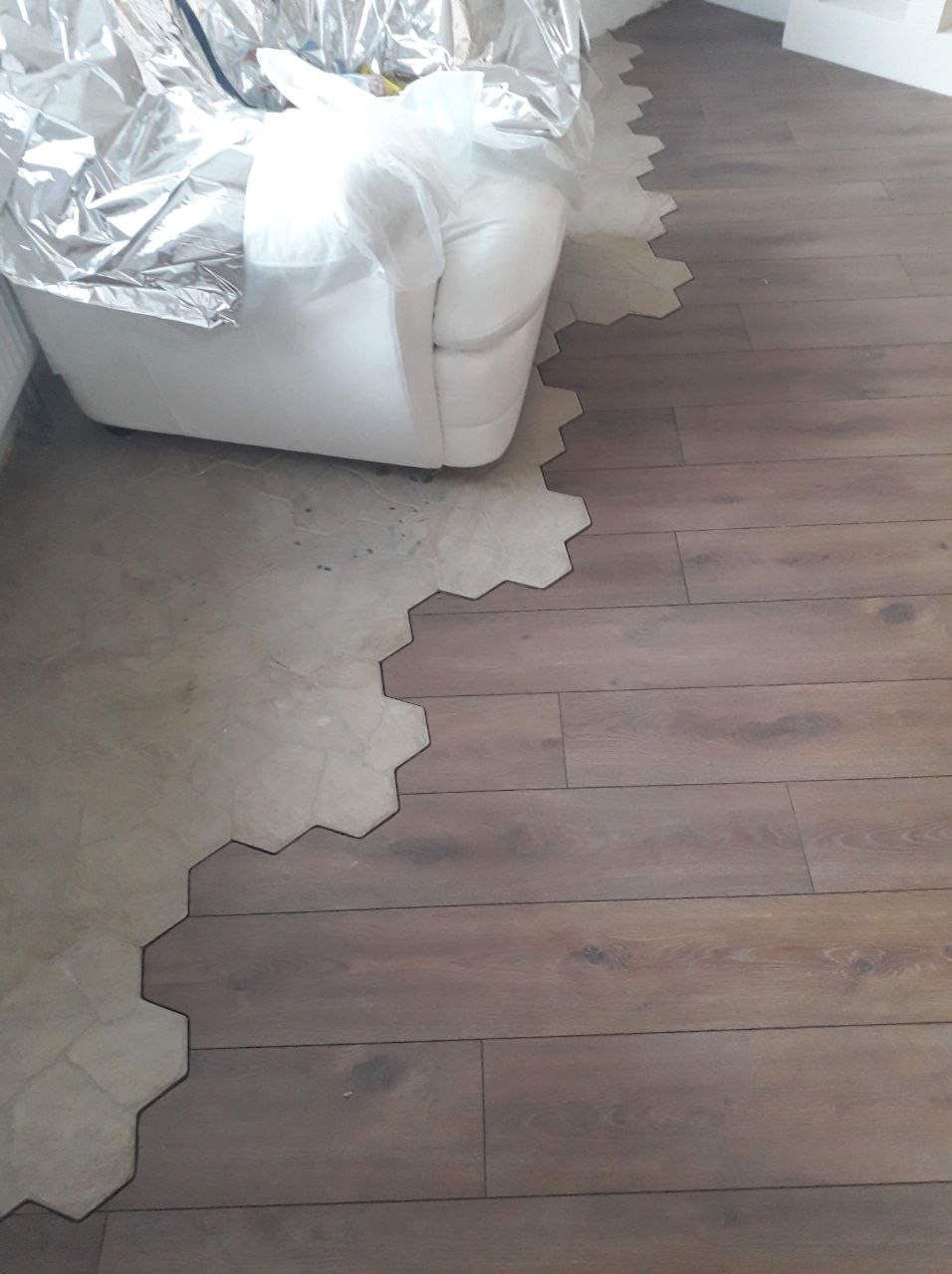 Fragment podłogi z brązowych paneli przechodzących w sześciokątne płytki, z meblem zabezpieczonym folią w tle.