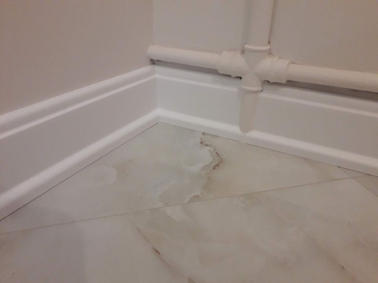 Fragment wnętrza: białe listwy przypodłogowe, marmuropodobna podłoga z brązowymi żyłkami i rura grzewcza w rogu pomieszczenia.