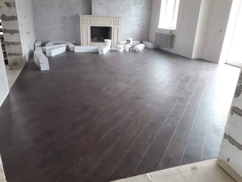 Ciemnobrązowa podłoga ułożona w jodełkę w przestronnym pokoju z kominkiem i grzejnikiem, częściowo zakryta paczkami materiałów budowlanych i pojemnikami z klejem.