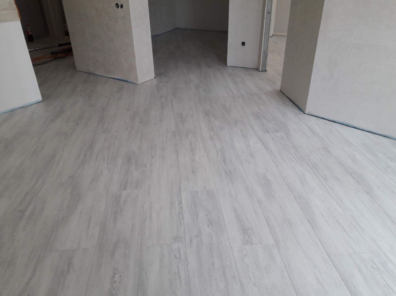 Jasnoszara podłoga z paneli imitujących drewno w nowym, pustym pomieszczeniu z surowymi ścianami i widocznymi otworami w ścianach.