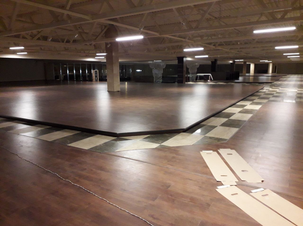 Rozległa sala z częściowo ułożoną, ciemnobrązową podłogą, widoczne panele w opakowaniach, wzór szachownicy na obrzeżach, oświetlenie sufitowe.