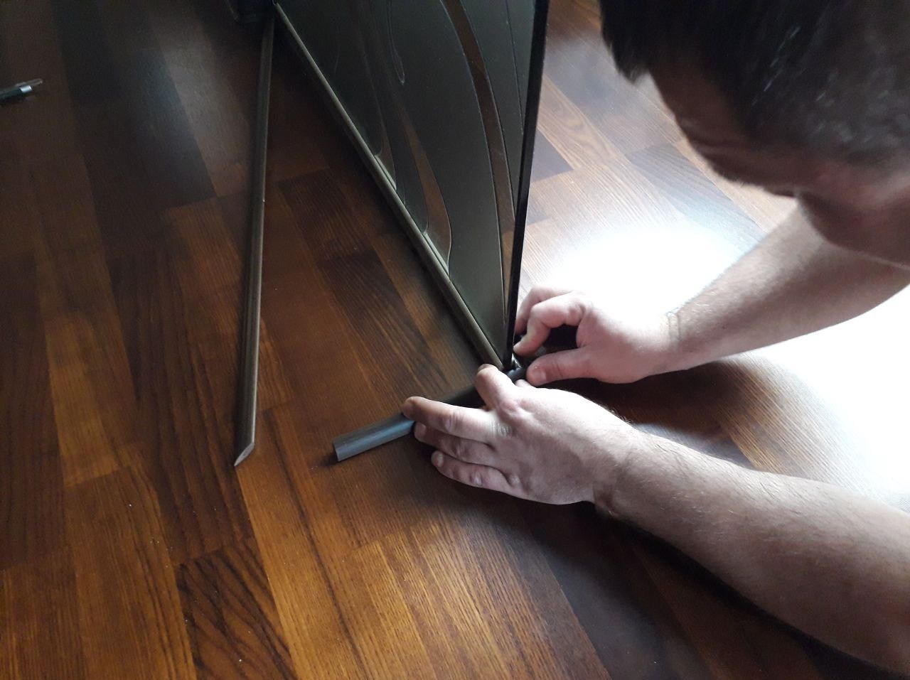 Pracownik montuje listwę przypodłogową przy ciemnym panelu podłogowym, widoczne dłonie i fragment narzędzia.