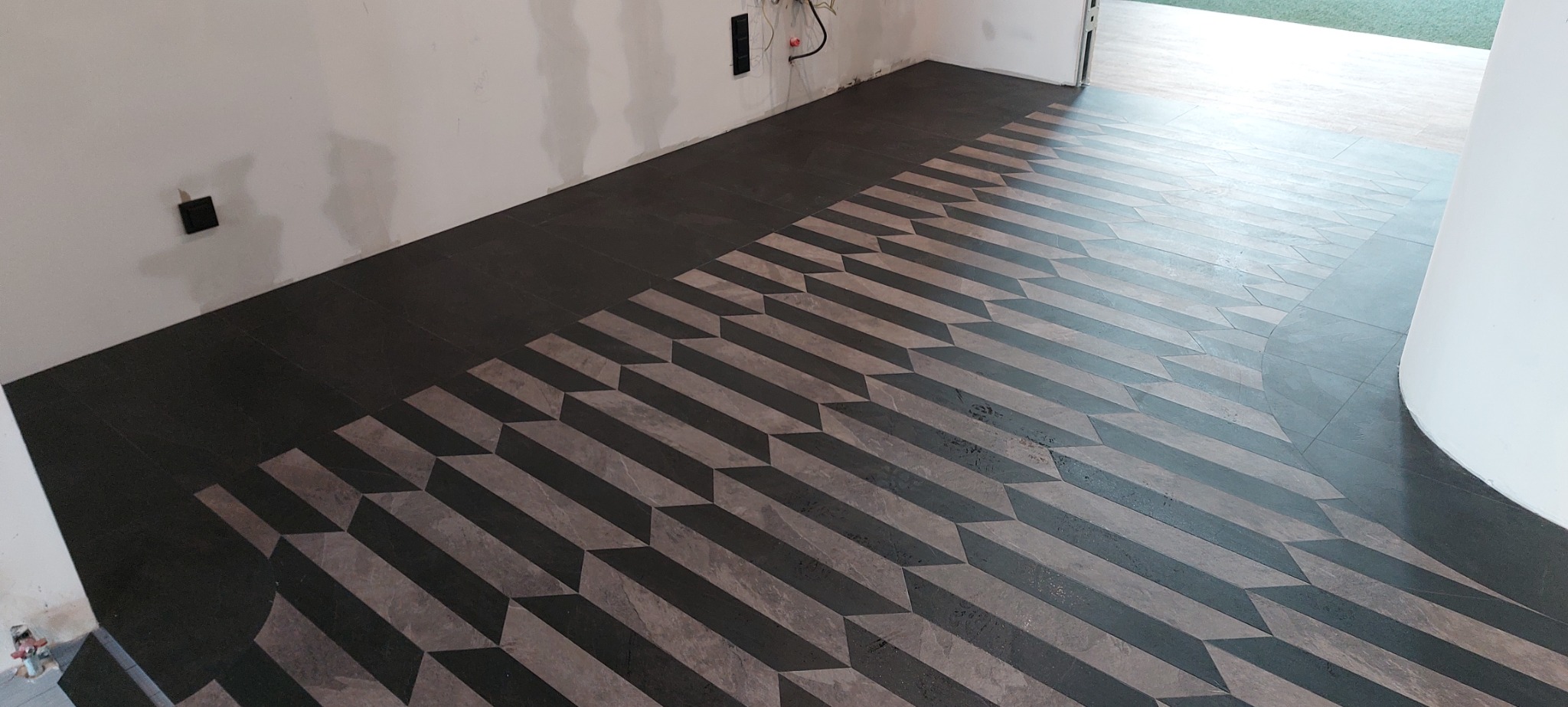 Ukończona podłoga z płytek ceramicznych ułożonych we wzór geometryczny 3D, ciemnoszare i jasnoszare płytki, widoczne gniazdka elektryczne na ścianie w tle.