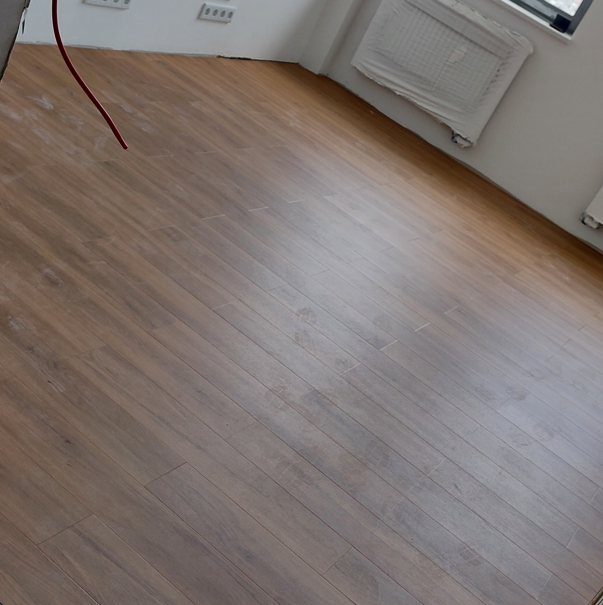 Układanie paneli podłogowych w jasnym pomieszczeniu, widoczne ślady po pracy, zabezpieczony grzejnik, wystający kabel.