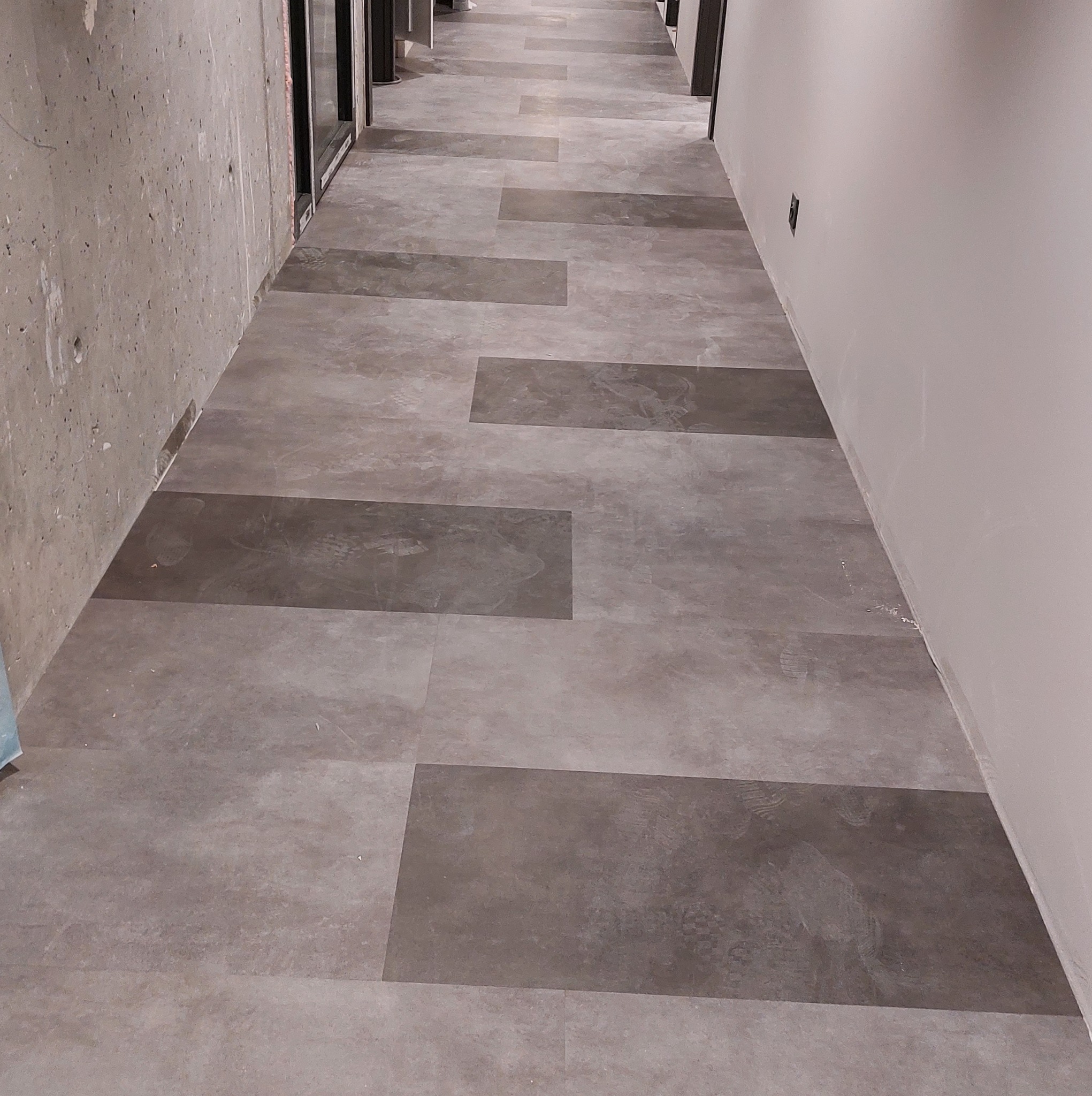 Korytarz z szarą podłogą wyłożoną prostokątnymi płytkami imitującymi kamień, widoczne ślady butów na powierzchni, surowa ściana z betonu architektonicznego po lewej, świeżo pomalowana ściana po...