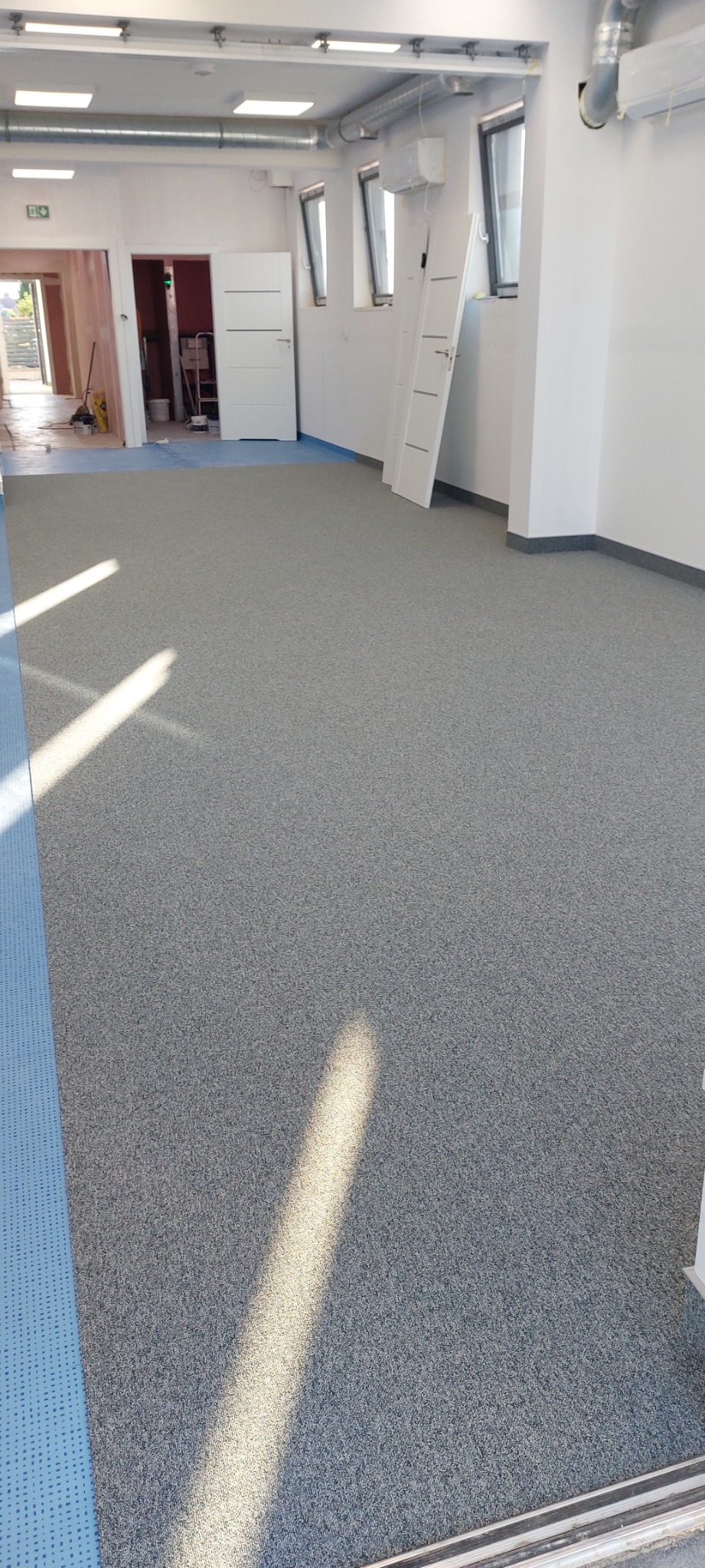 Szara wykładzina dywanowa w jasnym pomieszczeniu w Krakowie, widoczne fragmenty niebieskiej podłogi pod wykładziną, białe ściany z oknami i drzwiami, wpadające promienie słoneczne.