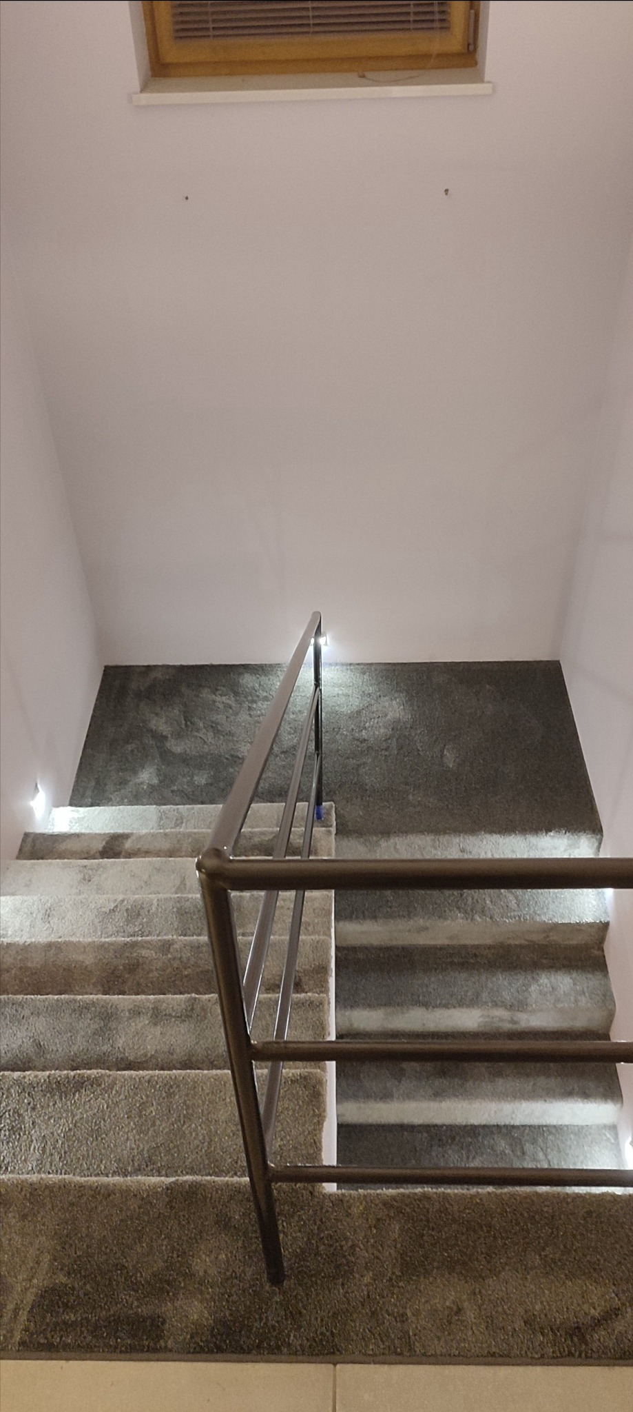 Widok z góry na schody wyłożone wykładziną dywanową w odcieniach szarości, z metalową balustradą i oświetleniem punktowym przy ścianie.
