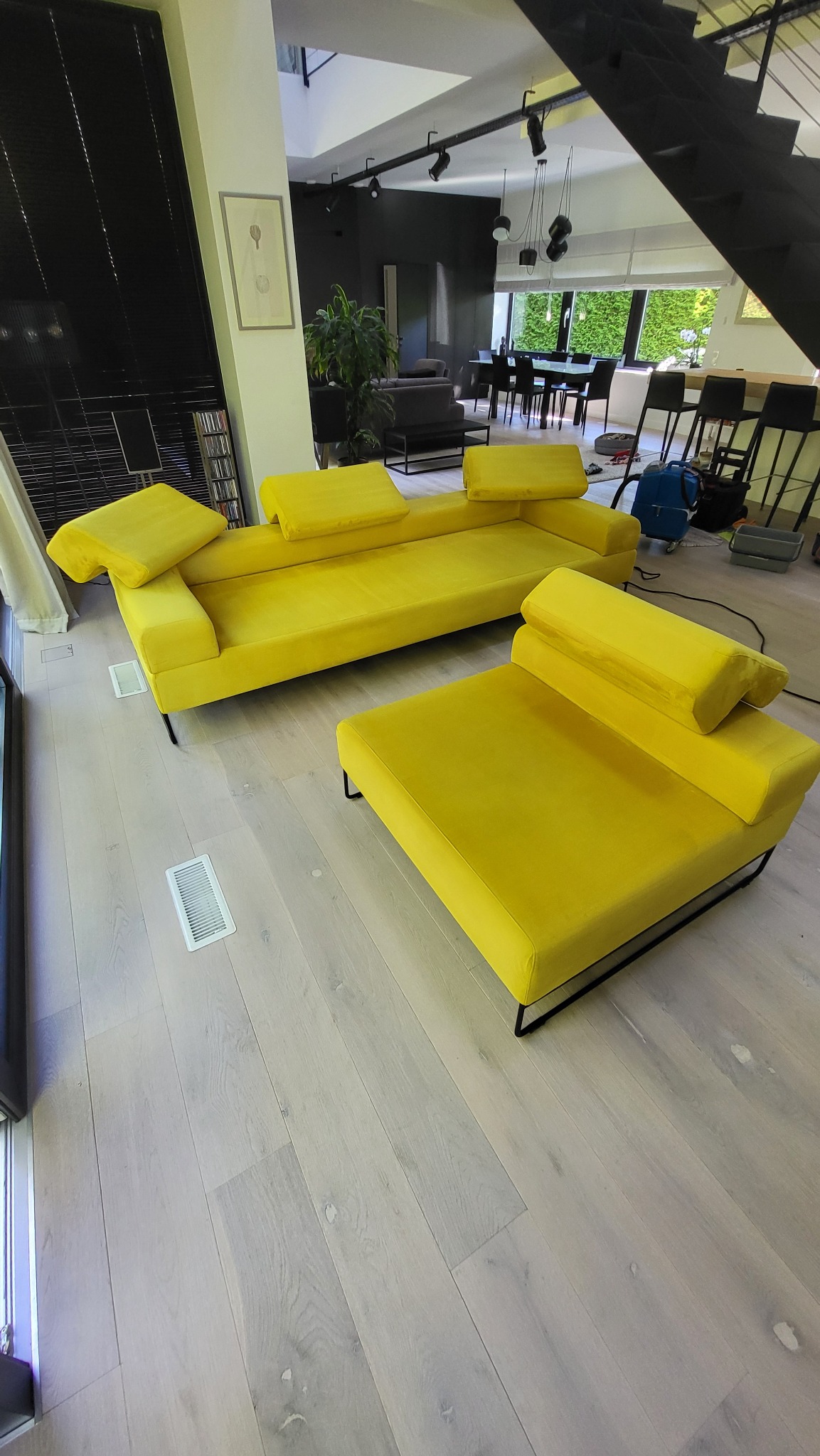 Nowoczesny, żółty zestaw wypoczynkowy z weluru, składający się z sofy i pufy, na jasnej drewnianej podłodze w przestronnym salonie z wysokim sufitem i schodami. W tle widoczny sprzęt czyszczący.