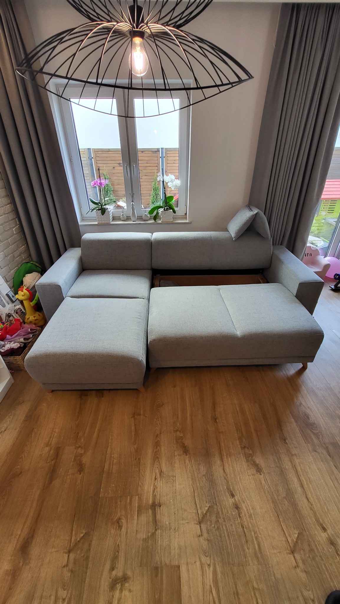 Rozłożona, modułowa, jasnoszara kanapa z funkcją spania i pojemnikiem na pościel, widoczna na drewnianej podłodze w pokoju z oknem i dekoracjami, oświetlona wiszącą lampą z żarówką Edison.