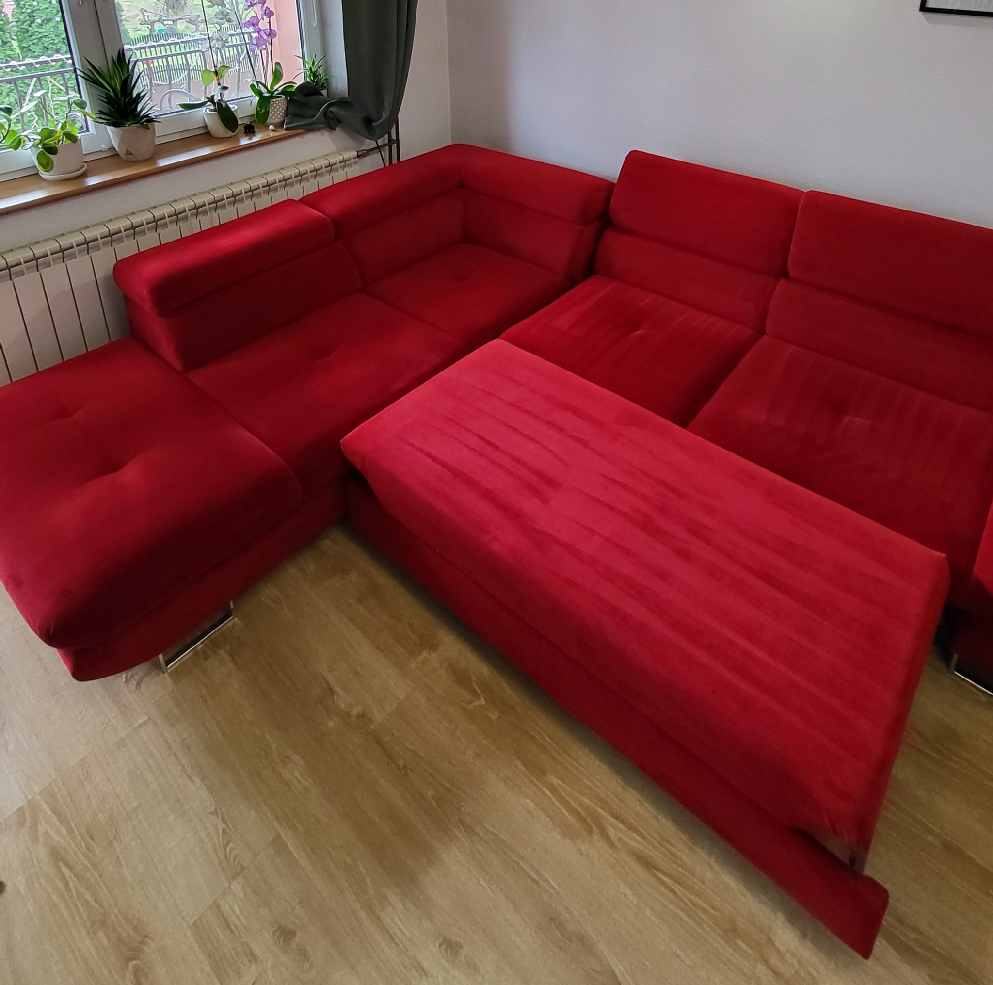 Czerwona, rozkładana sofa narożna z aksamitną tapicerką i wysuwanym podnóżkiem, widok z góry w jasnym wnętrzu.