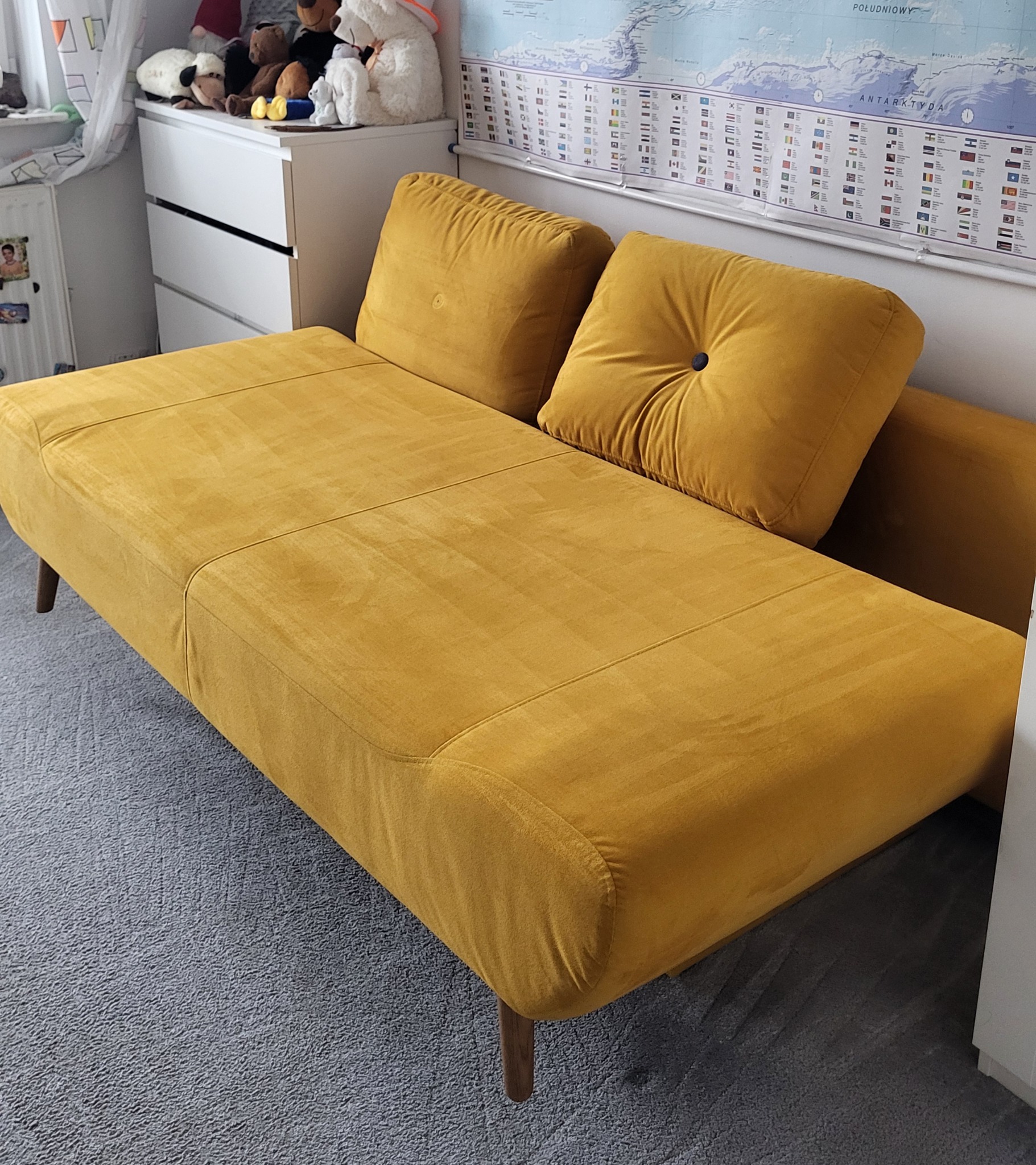 Musztardowa kanapa z dwoma poduszkami na tle dziecięcego pokoju z mapą świata i komodą z pluszakami.