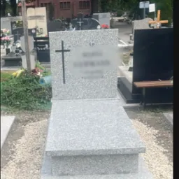 Jasnoszary, granitowy pomnik z krzyżem na cmentarzu, otoczony drobnymi kamykami, z rozmazanym napisem na tablicy. W tle inne nagrobki i budynek.