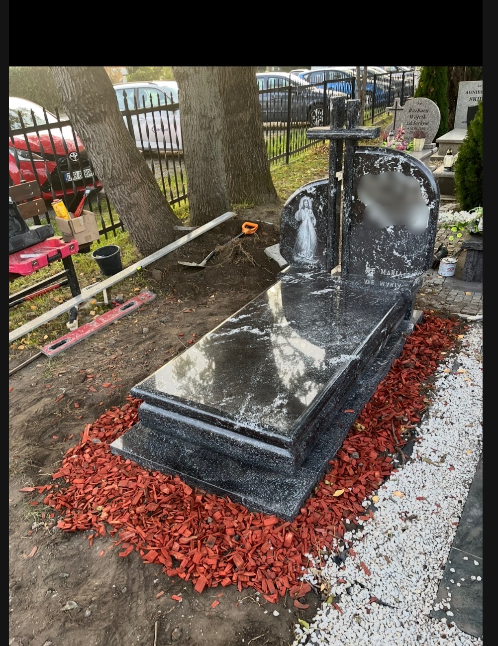 Czarny, polerowany pomnik z krzyżem i wizerunkiem postaci, otoczony czerwoną korą i białym grysem, na tle cmentarza z narzędziami do montażu w pobliżu.