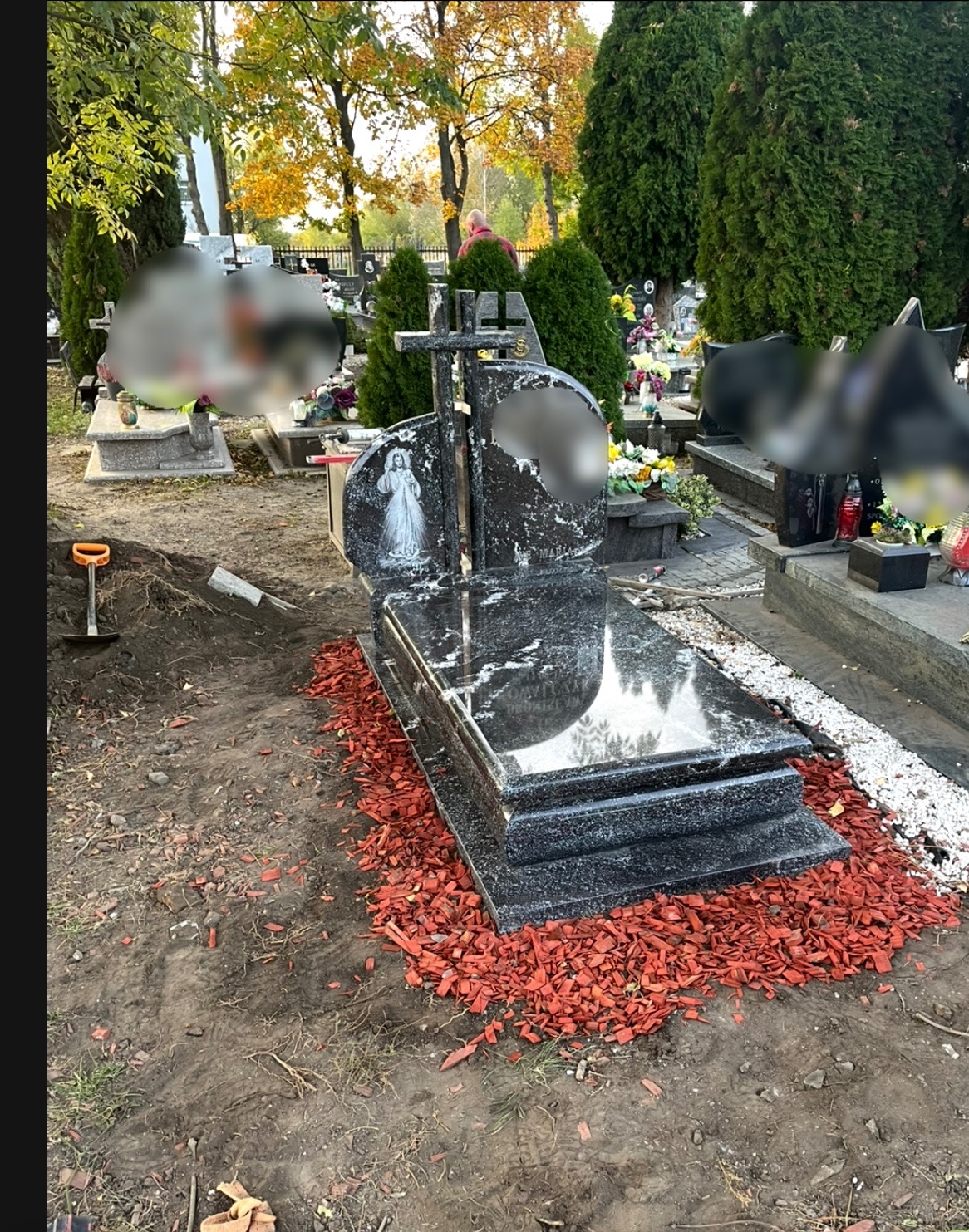 Nowy, czarny, polerowany nagrobek z wizerunkiem Matki Boskiej, otoczony czerwoną korą na cmentarzu, widoczna łopata wbita w ziemię.