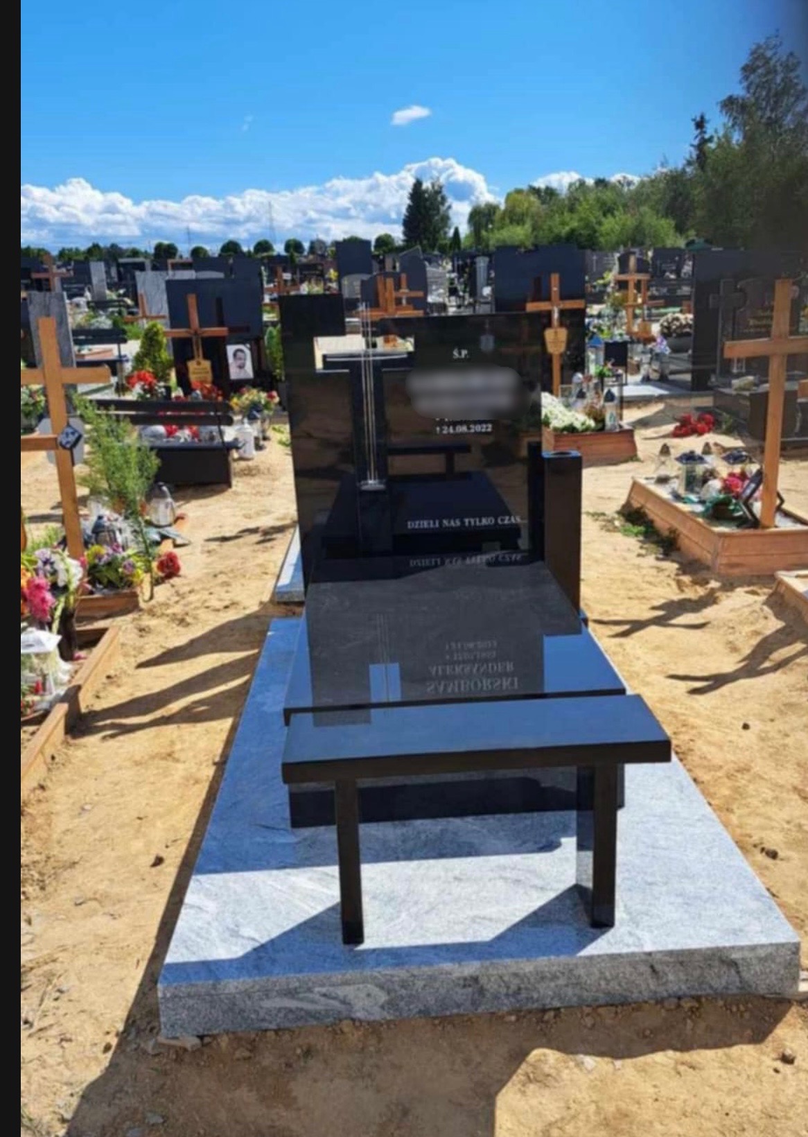 Czarny, nowoczesny pomnik z granitu z ławką na cmentarzu w słoneczny dzień, widok z przodu, na tle innych nagrobków.