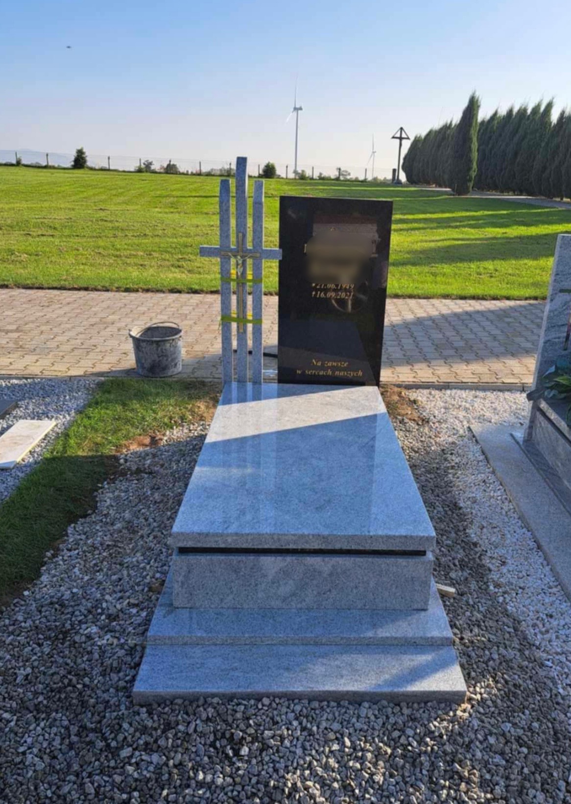 Granitowy pomnik z krzyżem w Stanowicach, na tle zielonego krajobrazu z turbinami wiatrowymi w oddali. Jasnoszara płyta nagrobna z ciemniejszą tablicą inskrypcyjną.