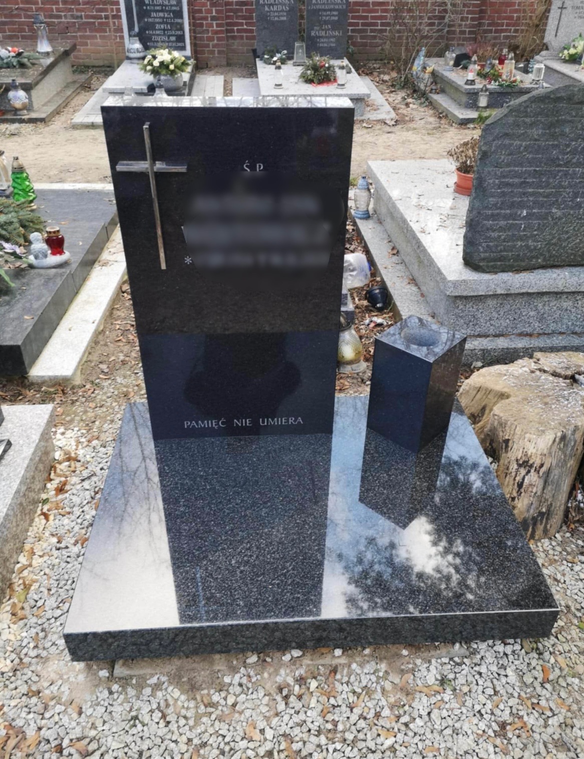 Czarny, polerowany pomnik z krzyżem i napisem 'Pamięć nie umiera' na cmentarzu, z widocznym odbiciem drzew w płycie podstawy.