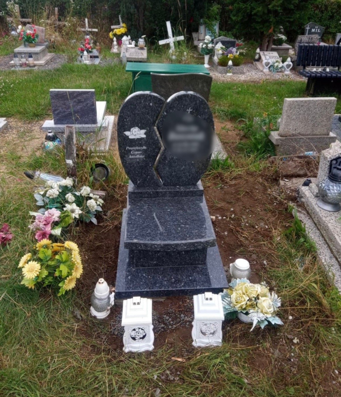 Granitowy pomnik w kształcie dwóch połączonych serc, z wygrawerowanym aniołkiem i inskrypcją, otoczony kwiatami i lampionami na cmentarzu.