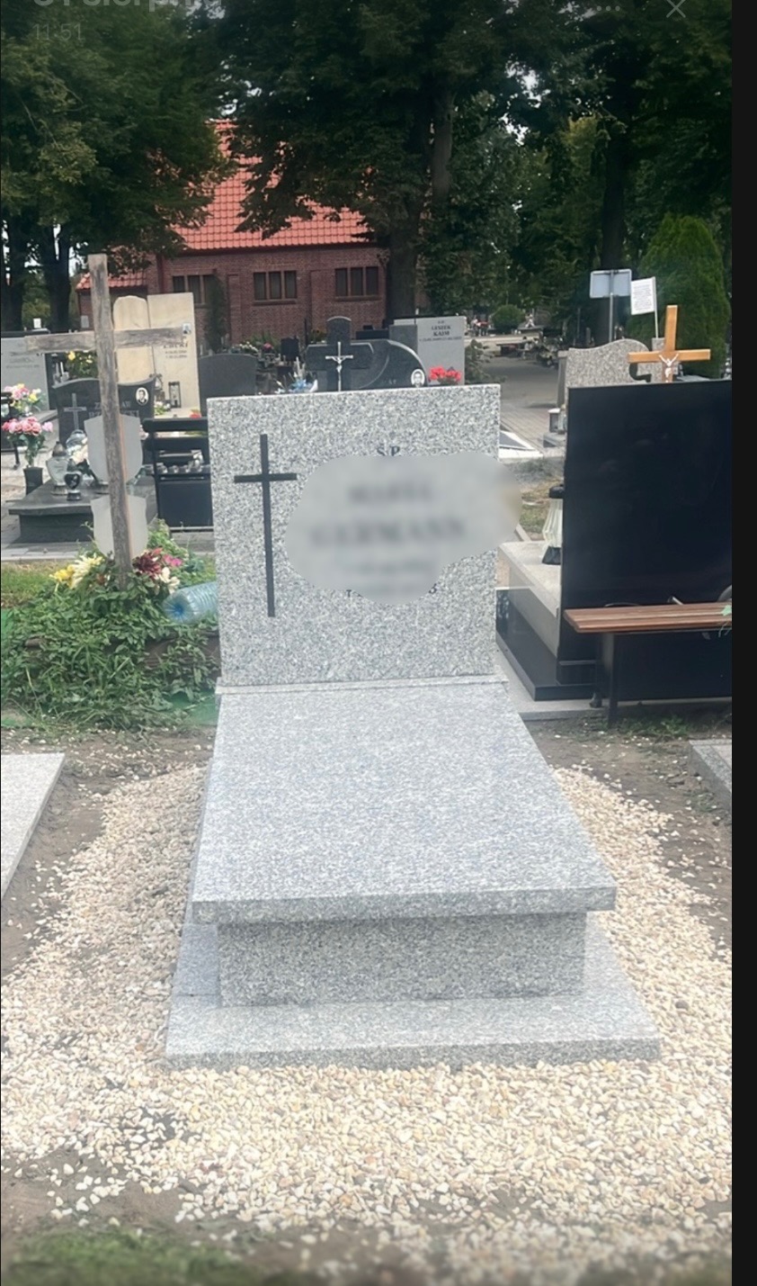 Jasnoszary, granitowy pomnik z krzyżem na cmentarzu, otoczony drobnymi kamykami, z rozmazanym napisem na tablicy. W tle inne nagrobki i budynek.