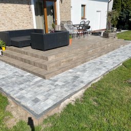 Romir-bruk - Szara kostka brukowa prowadzi do drewnianego tarasu z meblami ogrodowymi. Schody z płytek imitujących drewno. Trawa po bokach ścieżki. Słoneczny dzień.