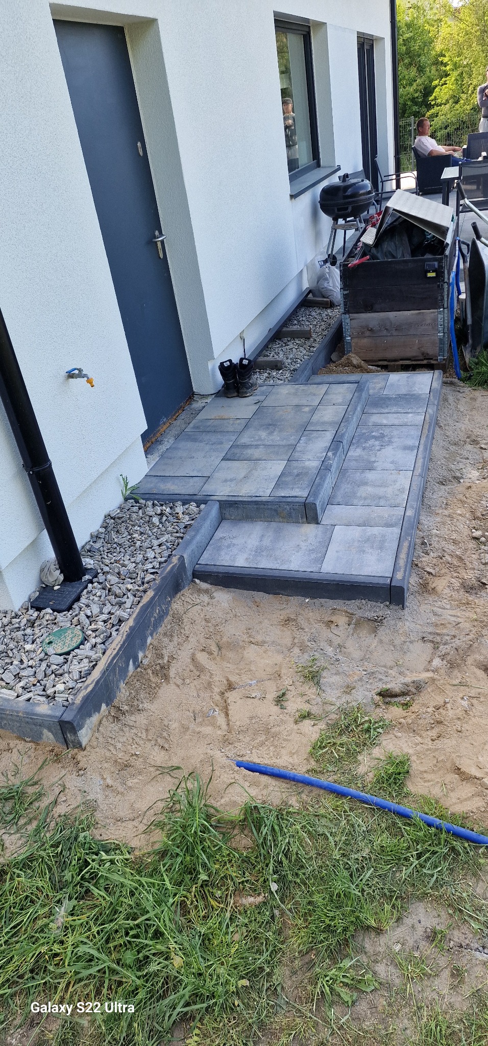 Schody z szarej kostki brukowej prowadzące do drzwi domu, obrzeża z kamyków, w tle grill i osoba siedząca na tarasie. Prace brukarskie w toku.
