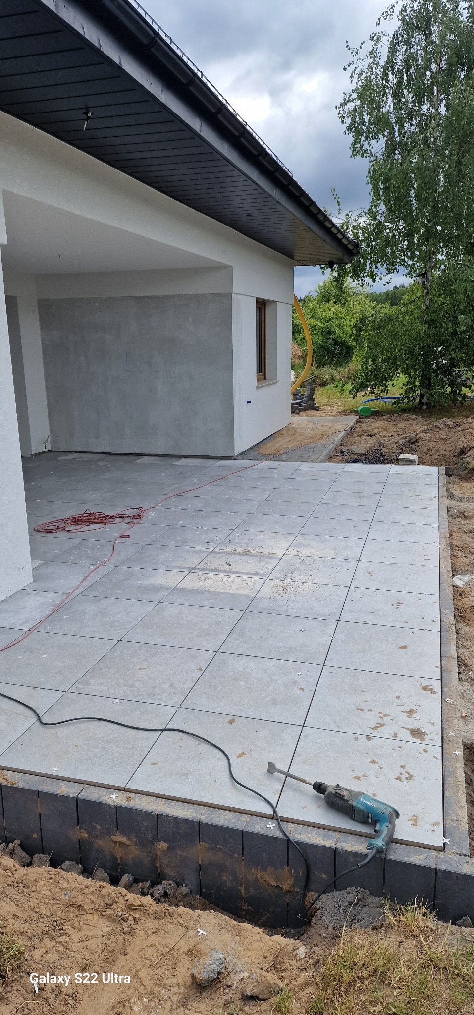 Jasnoszare płyty tarasowe ułożone przy budynku. Widoczny fragment elewacji i narzędzie pracy - wiertarka. Prace brukarskie w toku, zarys ogrodu w tle.