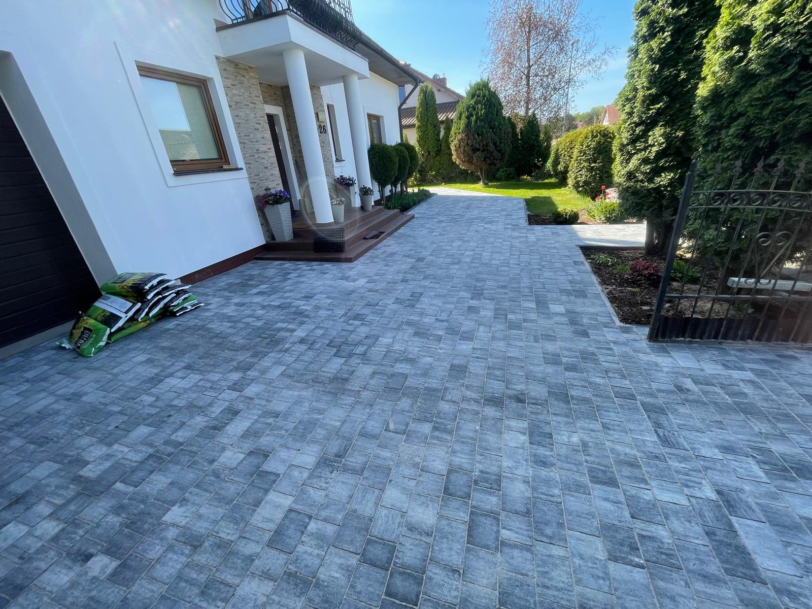 Nowoczesna, szara kostka brukowa prowadząca do domu z kolumnami. Stos worków z materiałem budowlanym obok. Słoneczny dzień, zadbany ogród.