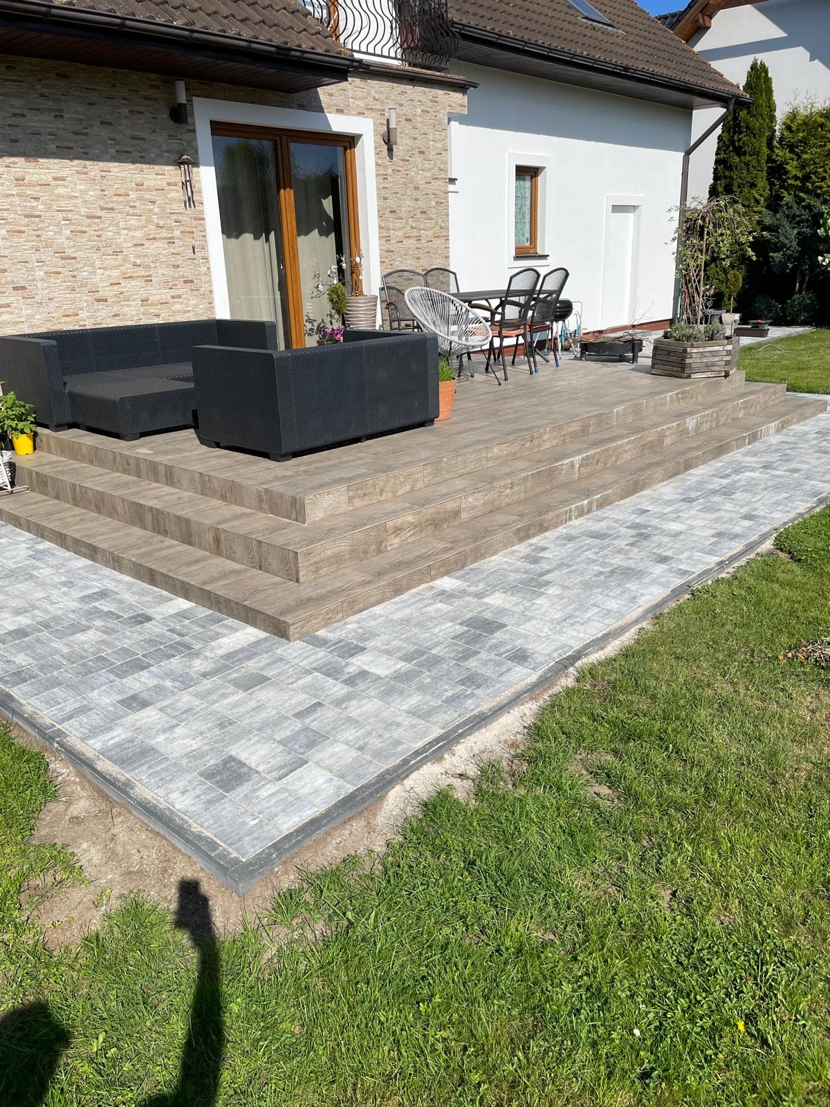 Szara kostka brukowa prowadzi do drewnianego tarasu z meblami ogrodowymi. Schody z płytek imitujących drewno. Trawa po bokach ścieżki. Słoneczny dzień.