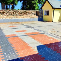 ZIBUD Usługi Elektryczno - Budowlane Zbigniew Ukleja - Wzór z czerwono-szarych kostek brukowych tworzący geometryczny deseń na podwórku z żółtą zabudową i kamiennym murem.
