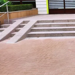 Wejście do budynku z rampą dla niepełnosprawnych i schodami, wykończone kostką brukową w ceglanym odcieniu, z elementami zieleni w tle i nowoczesną fasadą.