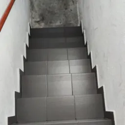 Schody wyłożone szarymi płytkami z poziomymi rowkami antypoślizgowymi, widok z góry, czerwona poręcz po lewej stronie, ściany w odcieniach bieli i szarości.