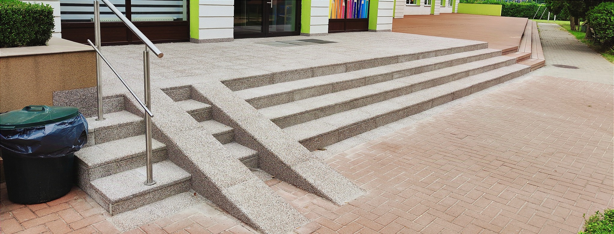 Zewnętrzne schody z podjazdami dla wózków, wykończone jasnym kamieniem dekoracyjnym, prowadzące do budynku z zielonymi akcentami, obok kosz na śmieci i chodnik z kostki brukowej.