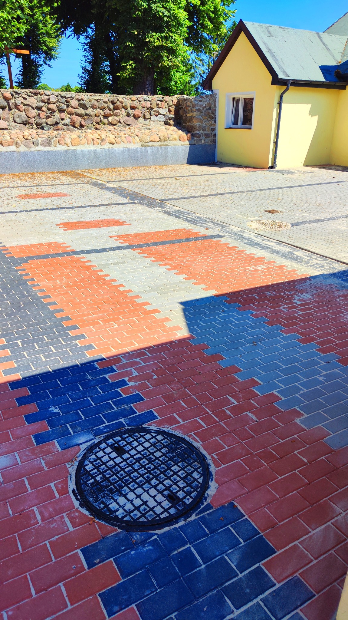 Kolorowa nawierzchnia z kostki brukowej w odcieniach czerwieni, szarości i granatu, ułożona we wzór, z okrągłą studzienką kanalizacyjną w dolnej części kadru, obok żółty budynek i kamienny murek.
