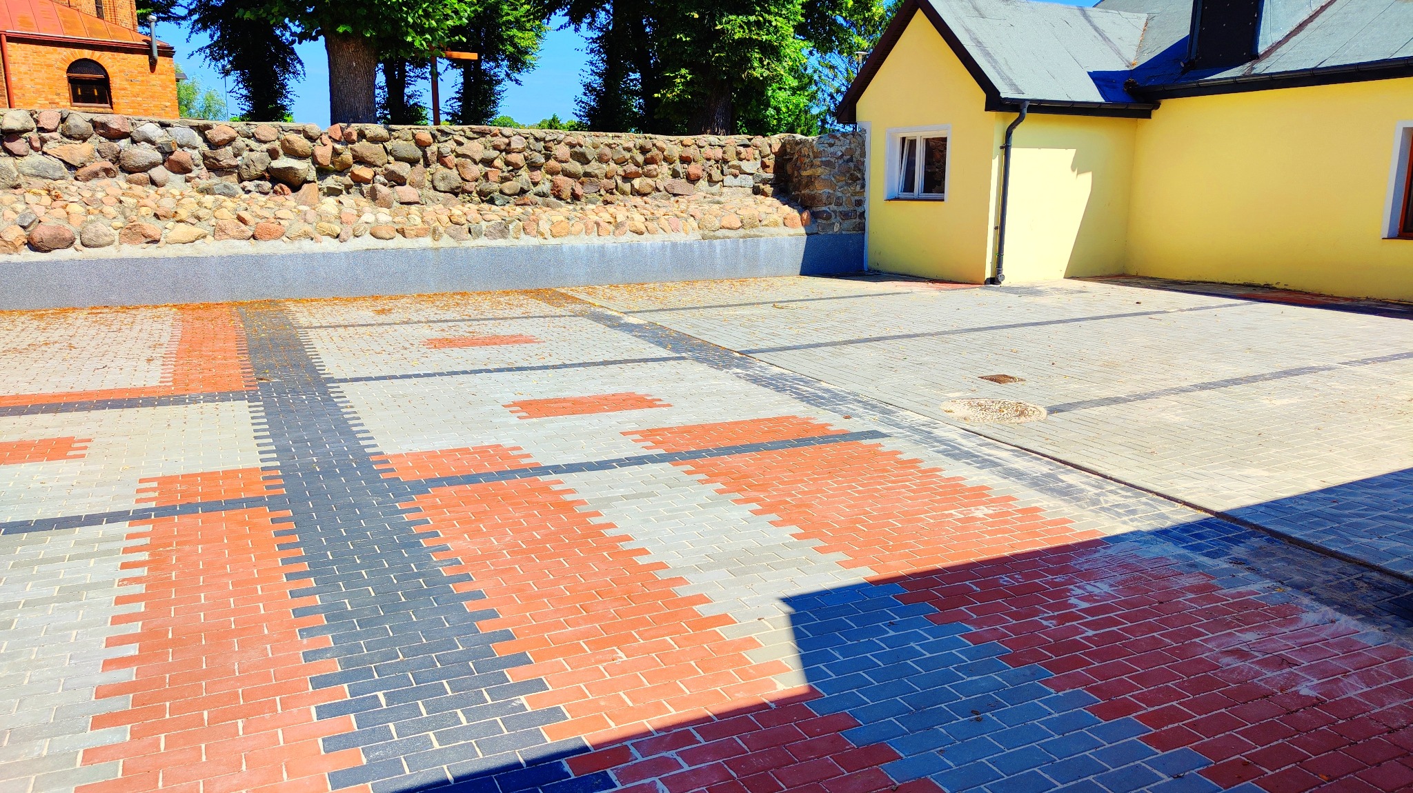 Wzór z czerwono-szarych kostek brukowych tworzący geometryczny deseń na podwórku z żółtą zabudową i kamiennym murem.