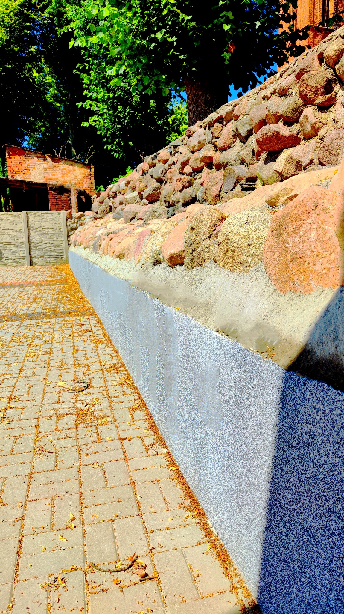 Kamienny murek oporowy z betonową obudową i kostką brukową, zasypaną opadłymi kwiatami lipy.